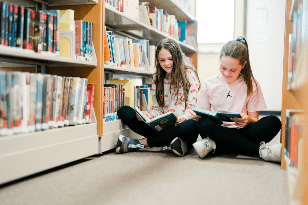 Two students sitting cross-legged on the floor in West Hills' library, reading the same book side by side.