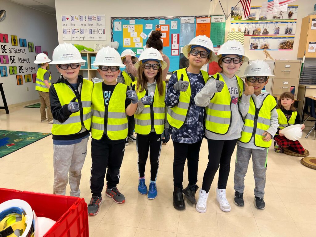 Young students dressed up like construction workers give a thumbs up to the camera.