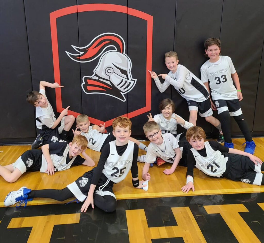 The elementary boys basketball team takes a picture with everyone pointing at the West Hills Knight Mascot Logo.