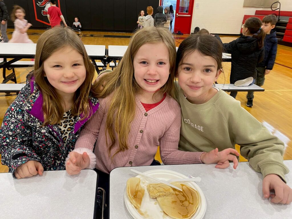 Three elementary girls smile and embrace each other for a photo at the lunch table at Portland area Christian School.