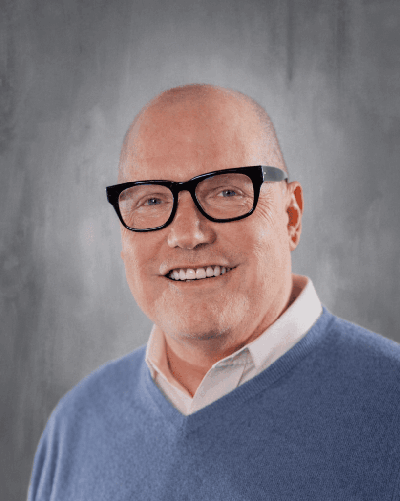 John Ogle smiling, wearing glasses and a blue sweater.
